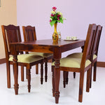 A solid wood dining table with four chairs arranged around it, the table has a vase with flowers on it and some plates and cutlery laid out, indicating a setting for a meal.