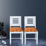 Two white finish solid wood dining chairs with red and orange upholstered cushions, placed against a dark background with a plant to the side.