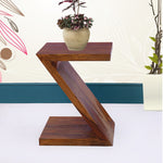 A wooden Z-shaped side end table in teak finish, with a vase of flowers on top, placed against a wall with decorative artwork.