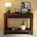 A solid wood console table with a drawer displayed in a living room setting, accompanied by decorative items such as a lamp, flowers, and framed picture.