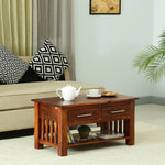 A rectangular solid wood coffee table with two drawers and a lower shelf, placed on a rug in a living room setting with decorative items on top.