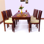 A solid wood dining table with a maple finish, accompanied by six chairs with beige upholstery. The table is set with a floral centerpiece and some decorative items.