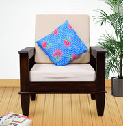 A single seater wooden sofa with a beige cushion and a blue decorative pillow with a floral design, placed in a room with a plant beside it.