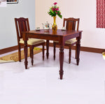 A wooden dining table with two chairs, featuring a maple finish, set in a room with a floral arrangement and tea set on the table.