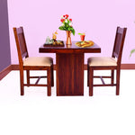 A solid wood dining table with two chairs set in a room with a purple wall, featuring a tablecloth, plates, cutlery, and a flower vase.