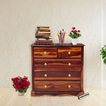 A wooden chest of drawers with honey finish, featuring multiple drawers with metal knobs, placed in a room with decorative items on top and flowers beside it.