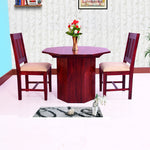 A dining table set with two chairs, the table has a dark wood finish and the chairs have a light beige fabric seats. The set is placed in a room with a light-colored wall and a floral arrangement on the table.