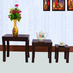 A set of three wooden nesting tables in walnut finish, decreasing in size from left to right, with a vase of flowers and a teapot on top of the largest table.