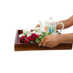 A wooden breakfast tray being held, with a cup, saucer, and a teapot on top of it, accompanied by a vase with red roses.