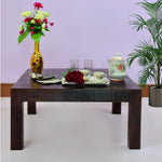 A square center coffee table made of walnut wood finish, with decorative items, a vase with flowers, and a tea set displayed on it. The table is placed against a purple backdrop with a potted plant to the right.