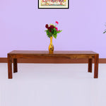 A wooden bench with a teak finish placed against a purple wall, with a vase of flowers on top.