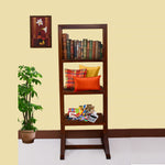 A wooden open bookshelf furniture with multiple items placed on its shelves, such as books, pillows, and decorative items. The bookshelf is positioned in a room with a plant and a framed picture on the wall in the background.