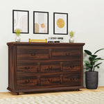 A wooden chest of drawers with multiple drawers, placed against a wall with decorative framed pictures, next to a potted plant. The chest has a rich wood finish and metal handles on the drawers.