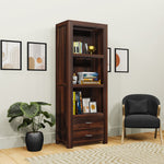 A wooden bookshelf with multiple shelves and a drawer, placed in a room with a chair and a potted plant, displaying a variety of books.