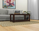 A solid wood coffee table with a glass top and a lower shelf, placed on a patterned rug. The table is brown in color and has a modern style.