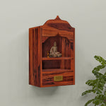 A wooden wall-mounted temple with a drawer, featuring a sculpture of a deity on the shelf, set against a white wall with a green plant beside it.