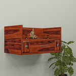 A wooden wall-mounted temple with a drawer, featuring a Buddha statue and LED lights, mounted on a wall beside a green plant.