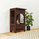 A wooden temple with a drawer, crafted in Sheesham wood with a mirror on the door, placed on a floor with a plant beside it.