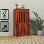 A wooden shoe rack with a drawer, finished in dark brown, placed against a wall with decorative items on top, situated in a bright room with a potted plant to the right.
