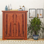 A wooden shoe rack with cabinet storage placed in a living room setting, featuring two brown doors and set against a wall with a decorative plant to the right.