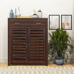 A brown solid wood shoe rack with a drawer, placed in a living room setting with decorative items on top and a potted plant beside it.