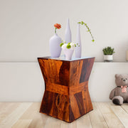 A wooden side table in a honey finish, with a decorative vase and Assorted plants on top, placed in a room setting with a toy on the side.