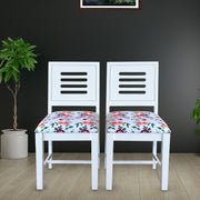 Two white wooden dining chairs with a floral pattern on the seat cushions, positioned in a room with a dark wall and a plant to the side.