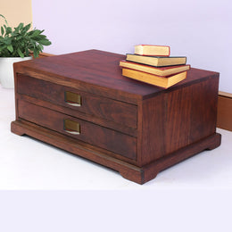 A brown Babool wood storage box with drawers, placed on a white floor against a wall, with a few books on top of the open drawer.