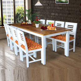 An 8 seater dining table set with solid sheesham wood with white finish accompanied by chairs with orange seat cushions, placed in a room with a brick wall and a window.