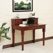 A wooden study table with drawers in a honey finish, placed against a wall with a framed picture above it. The table is hosting a laptop and has books and a plant on it.