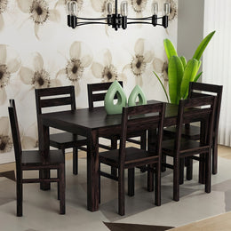 A wooden 6 seater dining table with chairs in a walnut finish, set against a background with floral wallpaper and a potted plant beside the table.