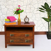 A solid wood shoe rack with two drawers, featuring metal handles, placed against a wall with decorative wallpaper. The shoe rack is arranged with a vase of flowers and a clock on top, next to a potted plant.