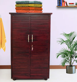 A brown solid wood wardrobe with two door and two drawer, with a plant and towels on top, set against a white wall.