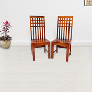 Two solid wood dining chairs with a honey finish, standing in a room with a white floor and wall, featuring a potted plant to the left.