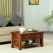 A rectangular solid wood coffee table with two drawers and a lower shelf, placed on a rug in a living room setting with decorative items on top.