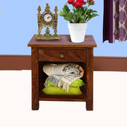 A wooden bedside table in walnut finish, featuring a storage compartment and a shelf with a vase of flowers on top, a clock on the left, and a pile of folded blankets on the shelf.