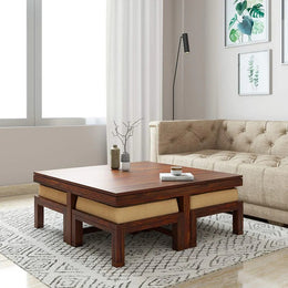 A wooden coffee table with beige cushions on four corners, placed on a white area rug in a living room setting, with a sofa in the background and a lamp on the wall.