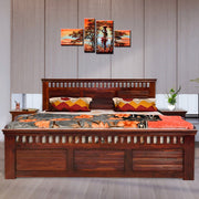A queen-size solid wood bed with storage drawers in a honey finish, accompanied by a set of four pillows with multicolored bedding. The bed is placed against a wall with a framed abstract painting above it.