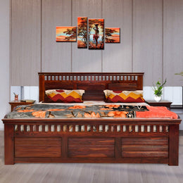 A queen-size solid wood bed with storage drawers in a honey finish, accompanied by a set of four pillows with multicolored bedding. The bed is placed against a wall with a framed abstract painting above it.