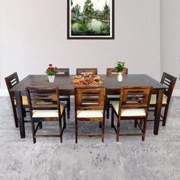 A rectangular wooden dining table with eight chairs around it, arranged for a meal. The room has a neutral color scheme, and there's a painting on the wall.