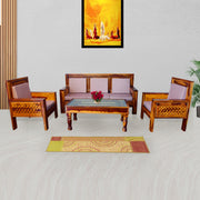 A brown wooden 5 seater sofa set arranged in a living room setting with a matching wooden table, the sofa set includes a three-seater sofa, a single seater sofa, and two armchairs, with a decorative rug and painting on the wall in the background.