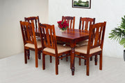 A wooden dining table with six chairs, the table and chairs are brown, the chairs have beige cushions, the room is well-lit with natural light and has a white floor.