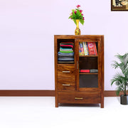 A brown finish solid wood standard bookcase with drawers, containing books and folded garments, with a vase of flowers on top.