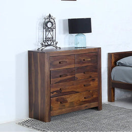 A brown wooden chest of drawers with a honey walnut finish, featuring five drawers, placed in a room next to a bed and decorative items on top.