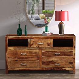 A rustic teak finish chest of drawers placed against a wall in a living room setting, featuring multiple drawers and open shelves, with decorative items on top.