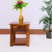 A wooden side end table in honey teak finish placed against a white wall with a yellow vase on top containing flowers. There is a potted plant beside the table.