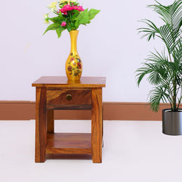 A wooden side end table in honey teak finish placed against a white wall with a yellow vase on top containing flowers. There is a potted plant beside the table.