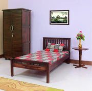 A solid wood single size bed with a checkered bedsheet, alongside a wooden armoire and a round wooden table with a vase of flowers, all placed in a room with purple walls and a framed picture on the wall.