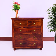 A solid wood chest of drawers with a maple finish, featuring five drawers and metal handles, placed in a room with a plant and a vase on top.