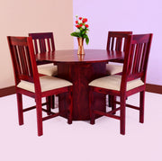 A round dining table with four chairs made of sheesham wood, featuring beige fabric cushions on the chairs. A vase with flowers is placed on the table.